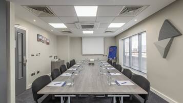 A conference room with a long table, chairs, a projector screen, and a window with blinds.