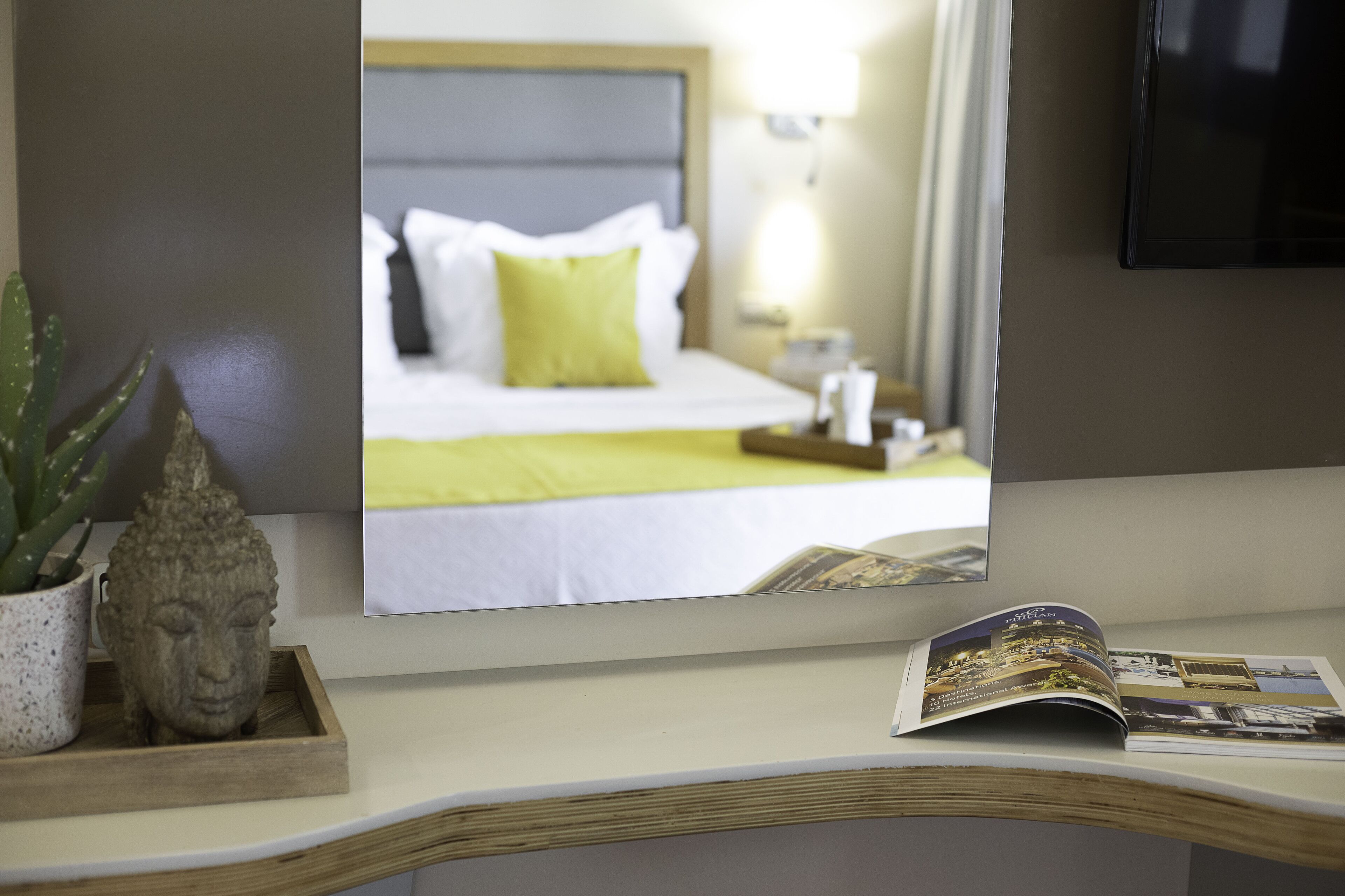 A hotel room with a bed, a nightstand, a lamp, a Buddha statue, and a magazine on a table.