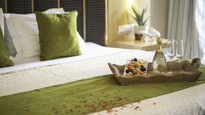 Un lit bien fait, agrémenté d’oreillers verts, un plateau avec des fruits et une bouteille de champagne, et des verres posés sur une table de chevet.