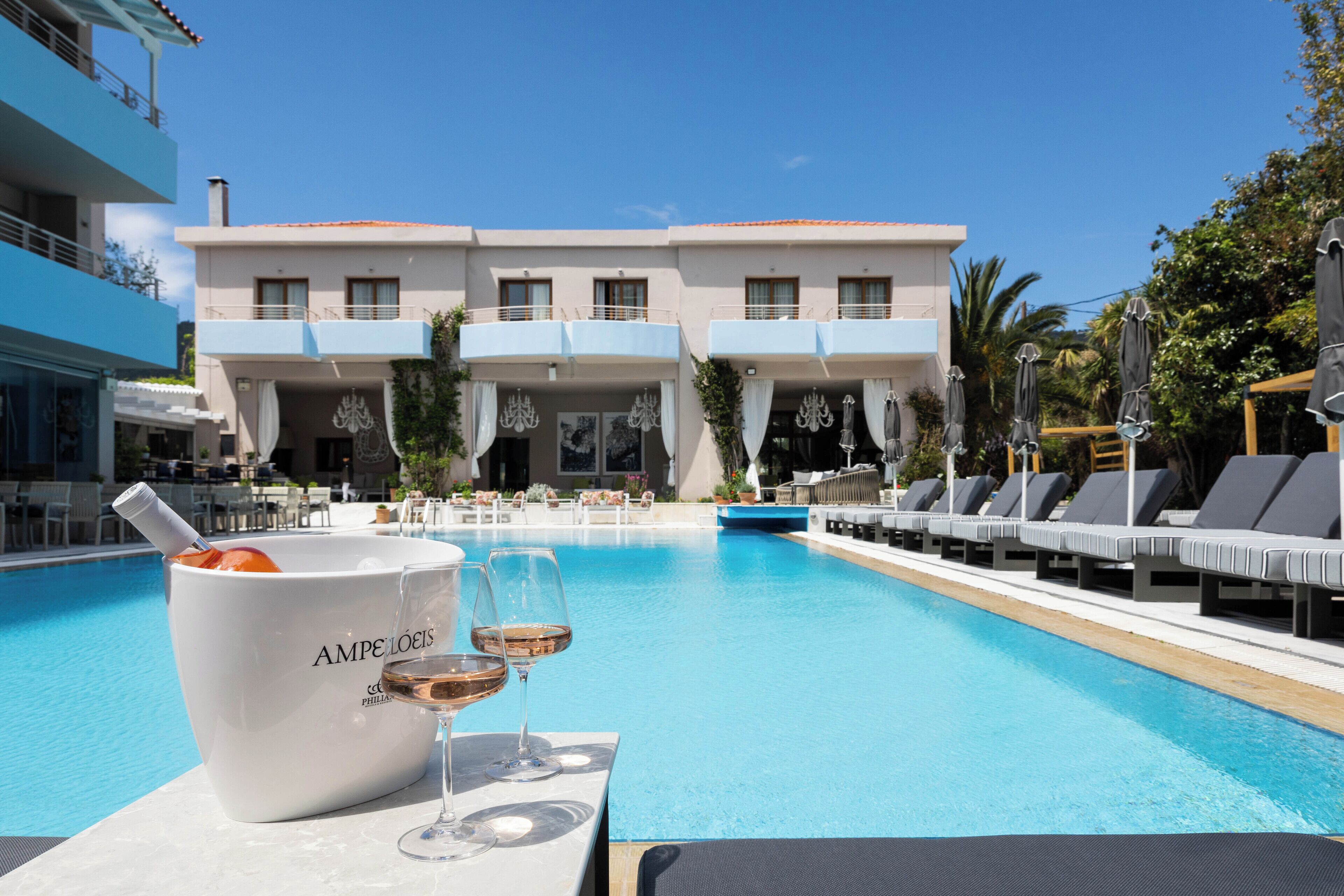 Área da piscina com espreguiçadeiras, um balde branco com a etiqueta 'AMPELLOIS' e dois copos de vinho.