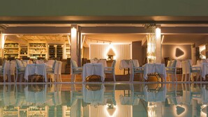 Un espace aménagé au bord de la piscine avec des tables et des chaises, un bar avec des étagères remplies de bouteilles, et le reflet de tout cela dans l’eau de la piscine.