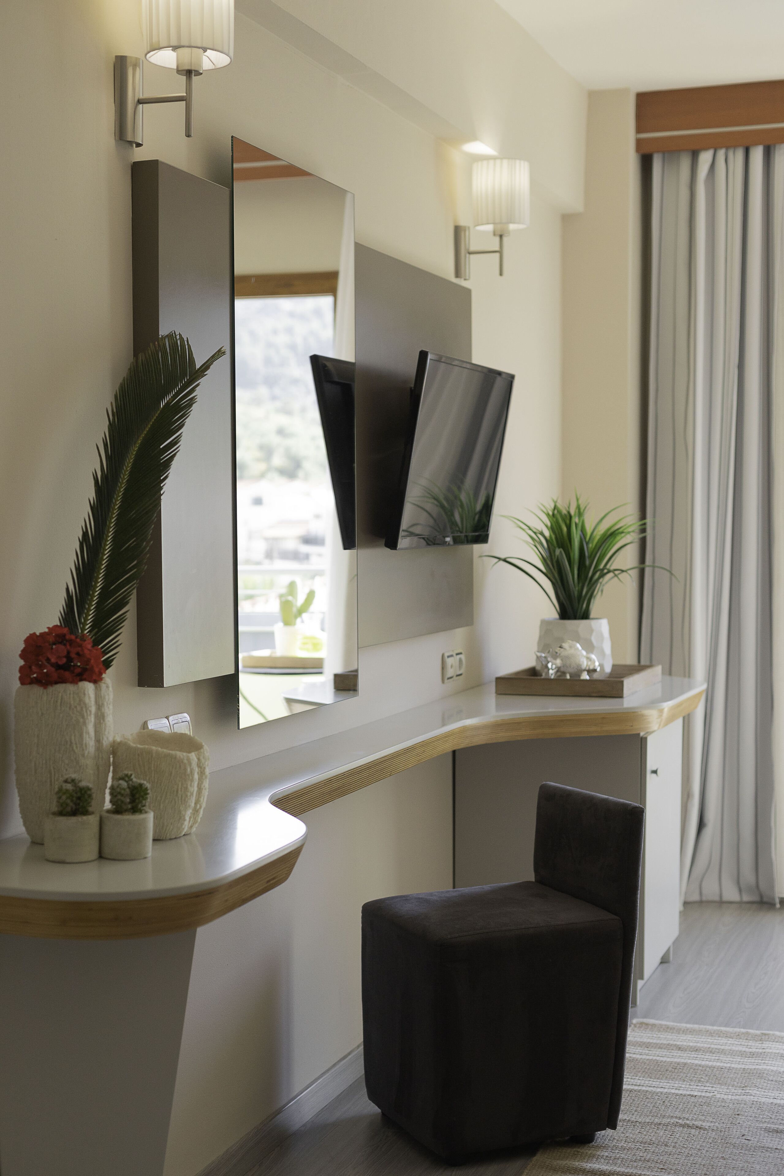 A modern hotel room with a wooden desk, a black ottoman, a large mirror, and wall-mounted lights.