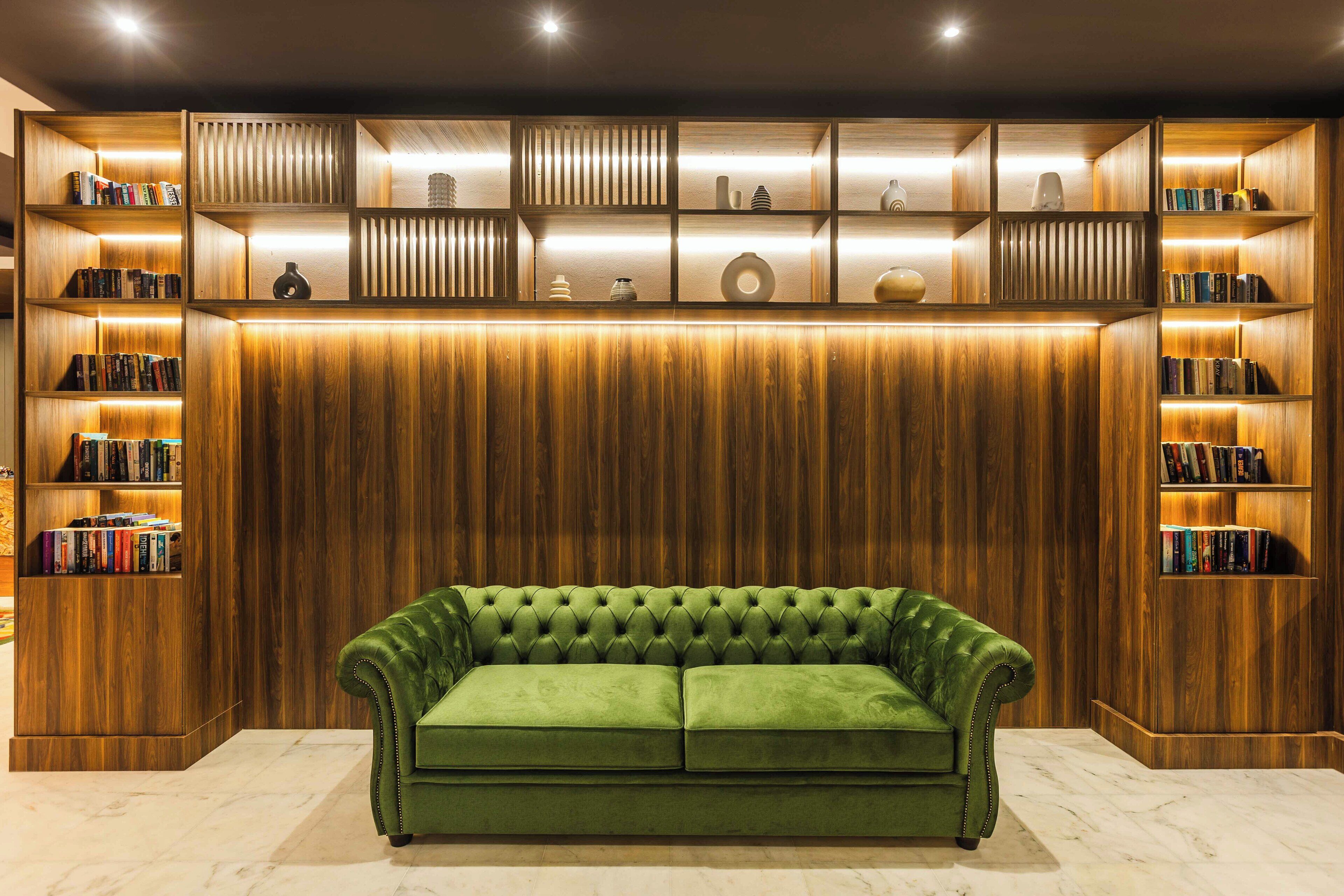 A green velvet sofa in front of a wooden bookshelf with illuminated shelves.