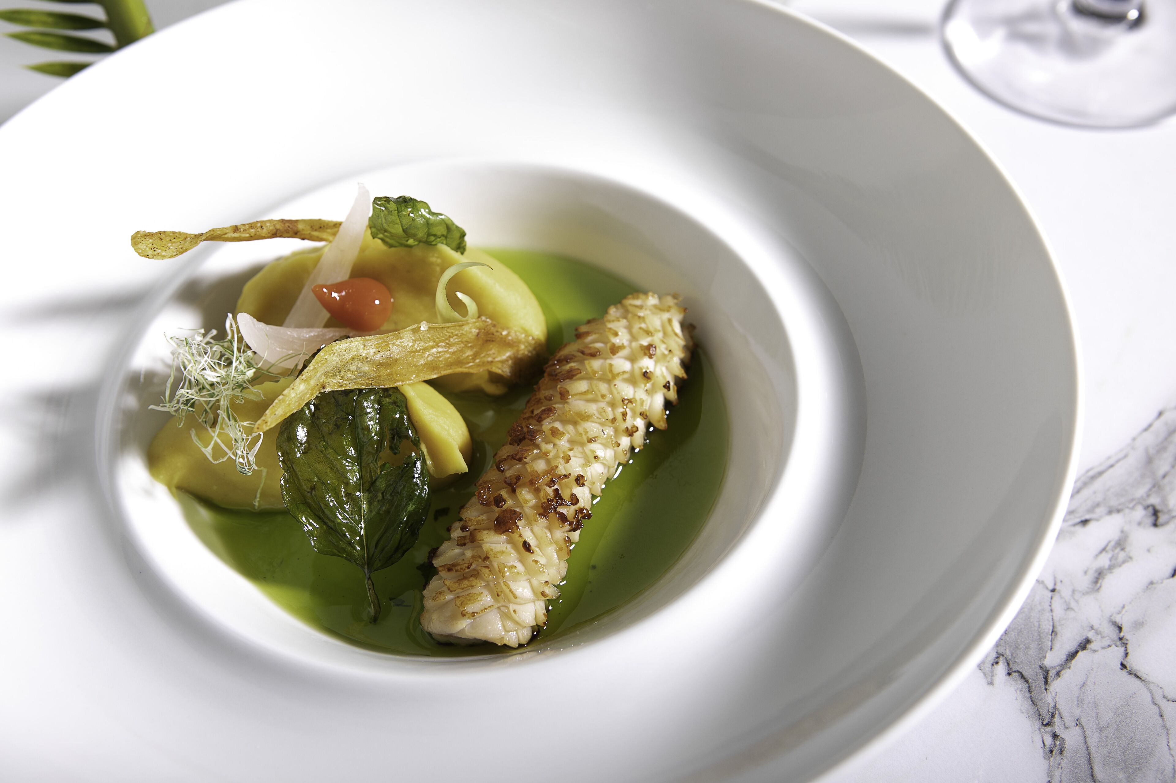 A white plate with a dish containing a piece of fish, vegetables, and a green sauce.