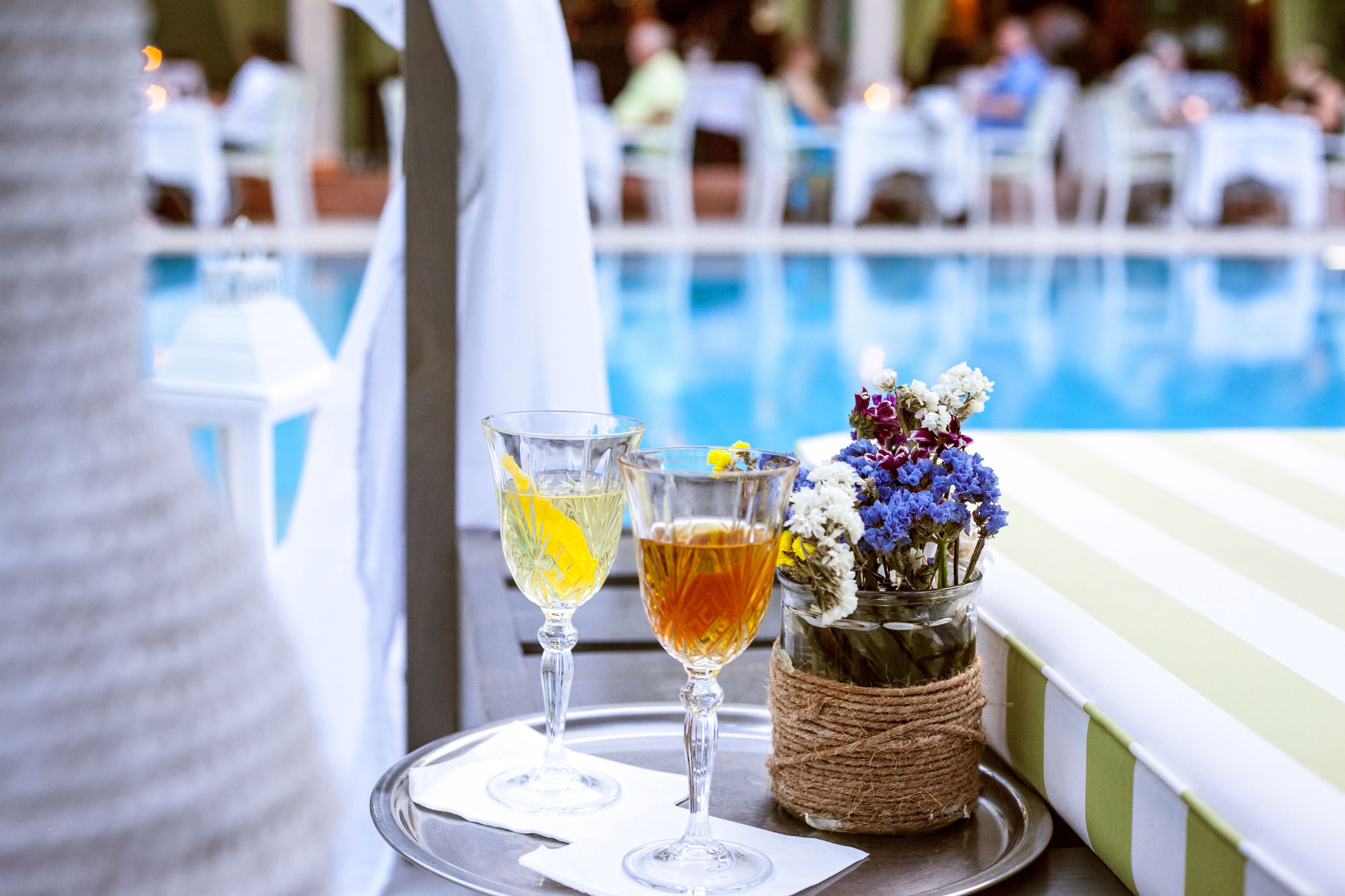 A poolside setting with two glasses of beverages on a tray, a vase with flowers, and a blurred background of people and tables.