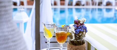 A poolside setting with two glasses of beverages on a tray, a vase with flowers, and a blurred background of people and tables.