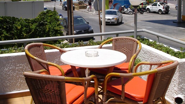 Un patio con sillas de mimbre y una mesa, con vista a una calle con coches y una gasolinera.