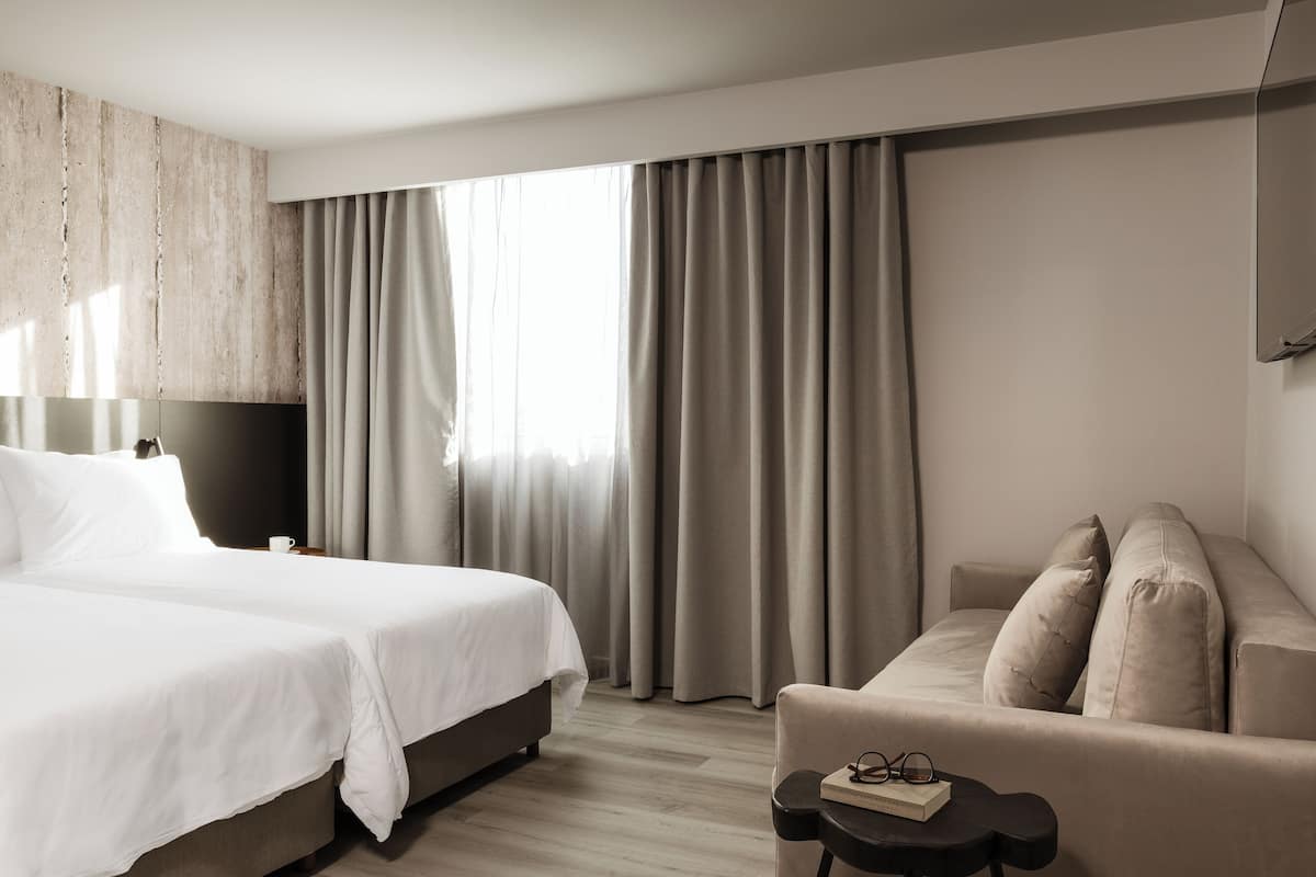 Habitación de hotel con una cama grande, un sofá, una mesita con un libro y vasos, y una ventana con cortinas.