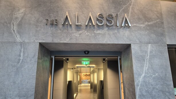 L’entrée de l’hôtel Alassia se distingue par son design moderne, avec une façade en marbre et un intérieur bien éclairé qui mène à un espace d’accueil.