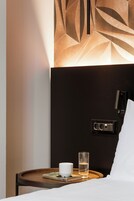 Uma cama bem arrumada com lençóis brancos, uma mesa de cabeceira com um livro, um copo de água e uma luminária na parede.