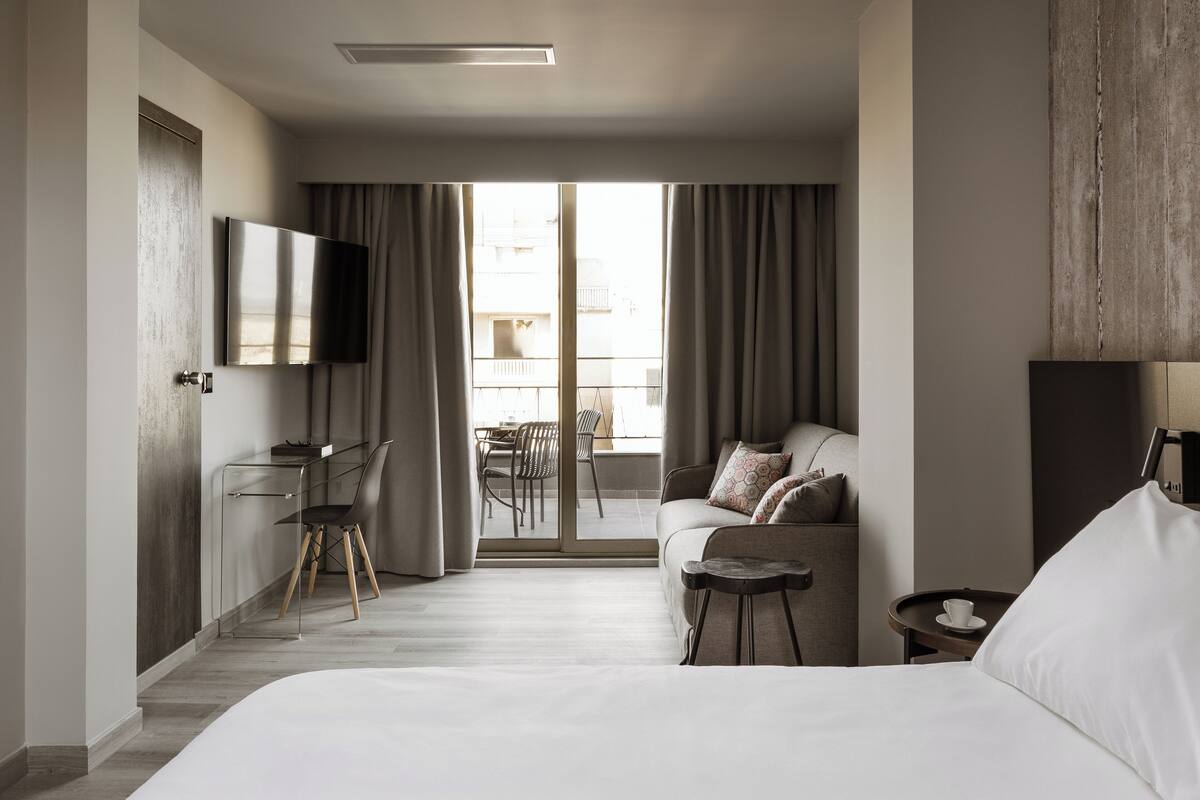 Una habitación de hotel moderna con cama, escritorio con silla, sofá, televisor montado en la pared y balcón con mesa y sillas.