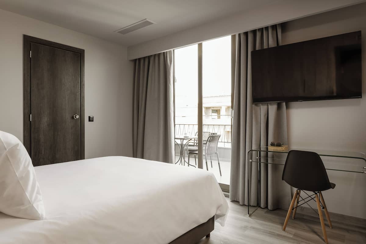 Habitación de hotel con cama, televisor montado en la pared, balcón con mesa y sillas, y una silla en la esquina.
