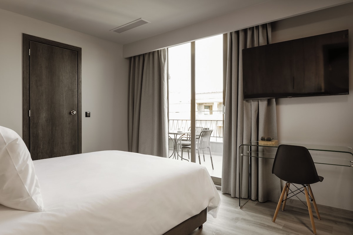A hotel room with a bed, a TV mounted on the wall, a balcony with a table and chairs, and a chair in the room.