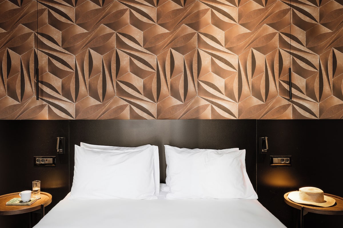 Una habitación de hotel con pared de diseño geométrico, una cama con ropa blanca, una mesita de noche con una taza y un sombrero, y una pequeña mesa redonda con una lámpara.