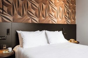 Un letto rifatto con lenzuola bianche, un comodino in legno con un bicchiere d'acqua e un libro, e un cappello appoggiato di lato.
