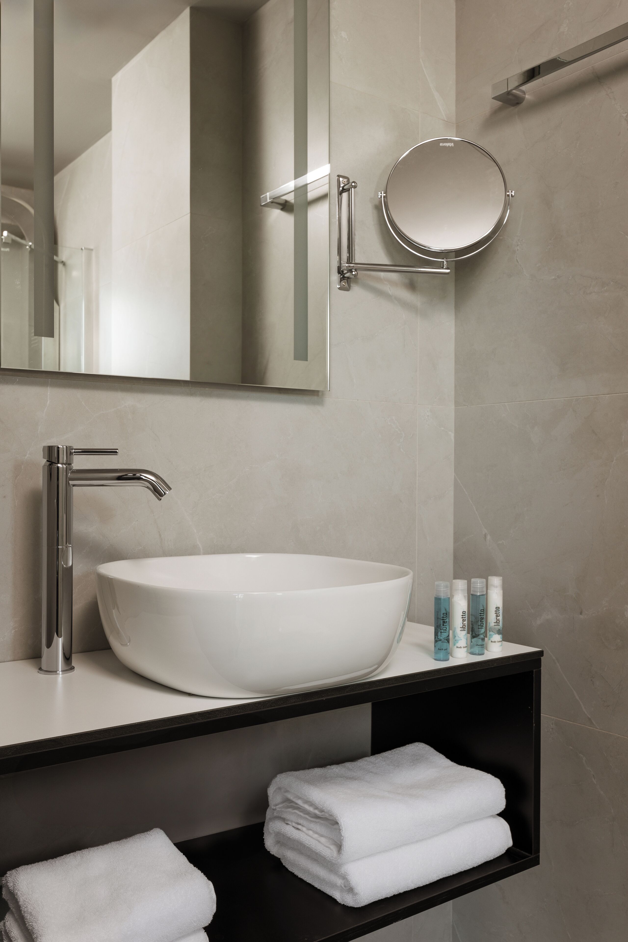 A modern bathroom with a large white sink, a round mirror, and a black shelf with towels.