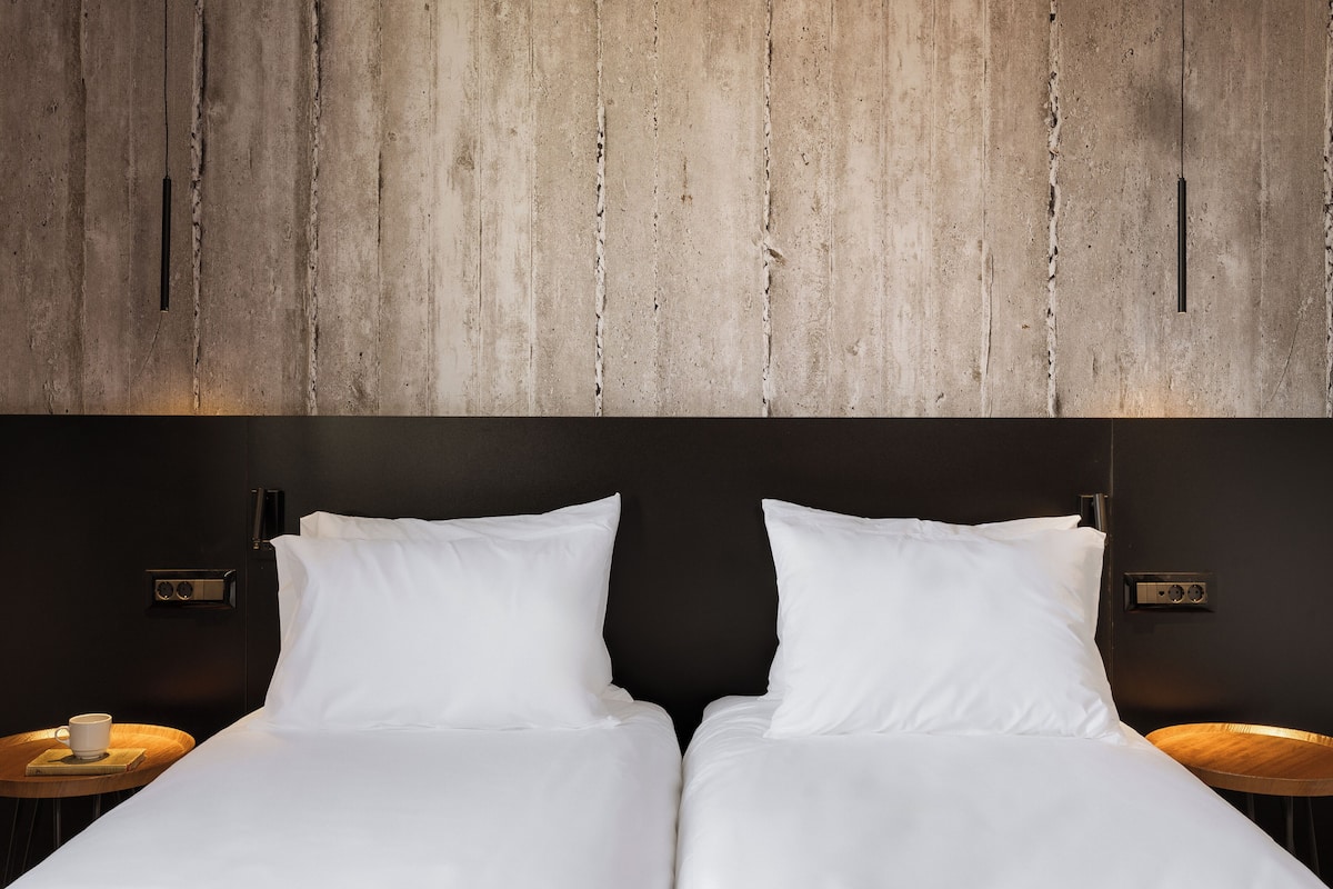 Una cama doble con sábanas blancas, una mesita de noche de madera con una taza, y una pared con diseño de paneles de madera.