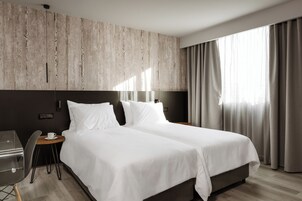 Uma cama de casal com lençóis brancos, uma mesa de cabeceira de madeira, uma pequena mesa com uma xícara e uma cadeira.