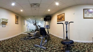 Gimnasio de hotel con equipo de ejercicio, piso alfombrado y obras de arte en las paredes.