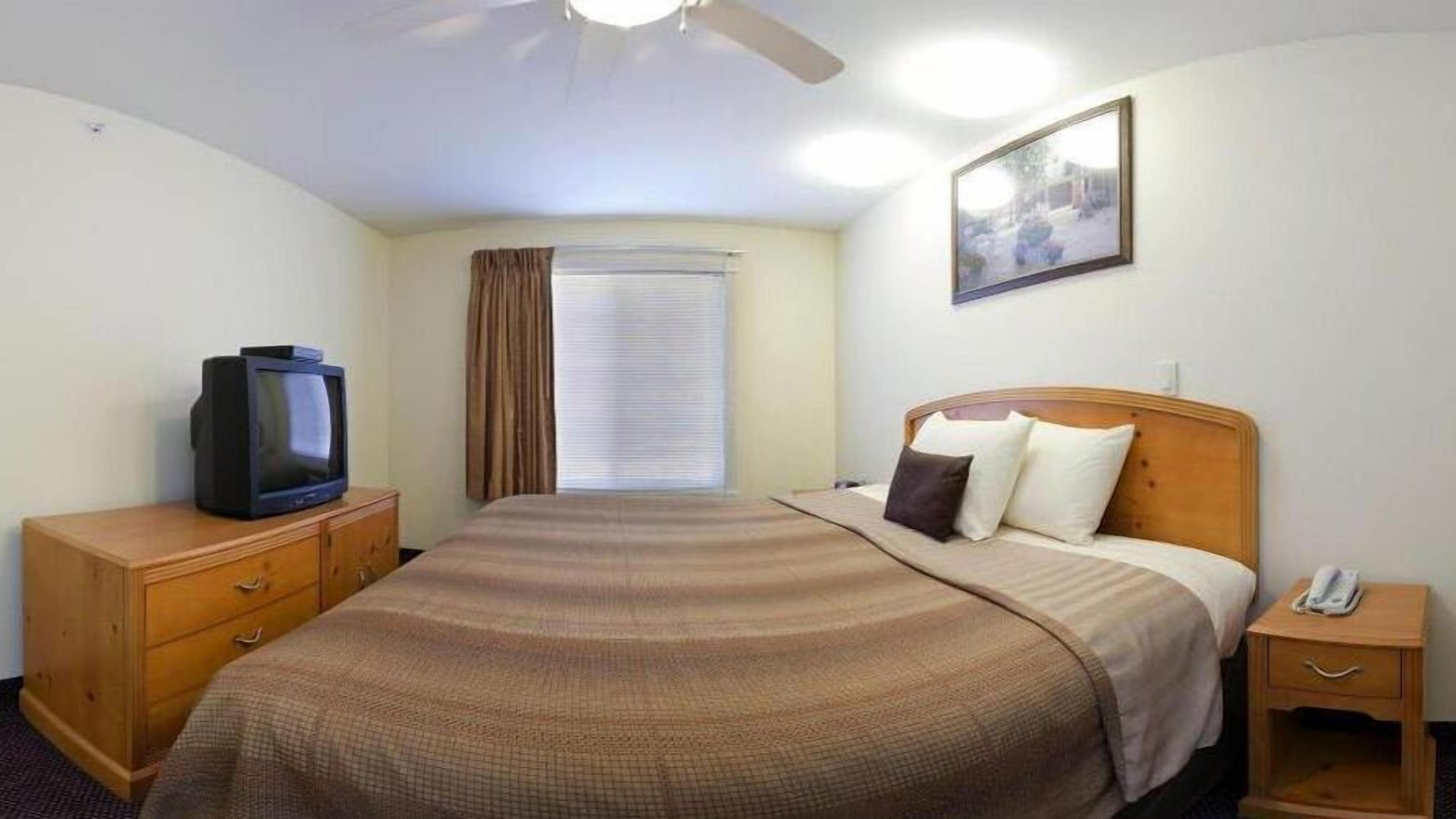 A hotel room with a bed, a television on a wooden dresser, a bedside table with a phone, and a window with curtains.