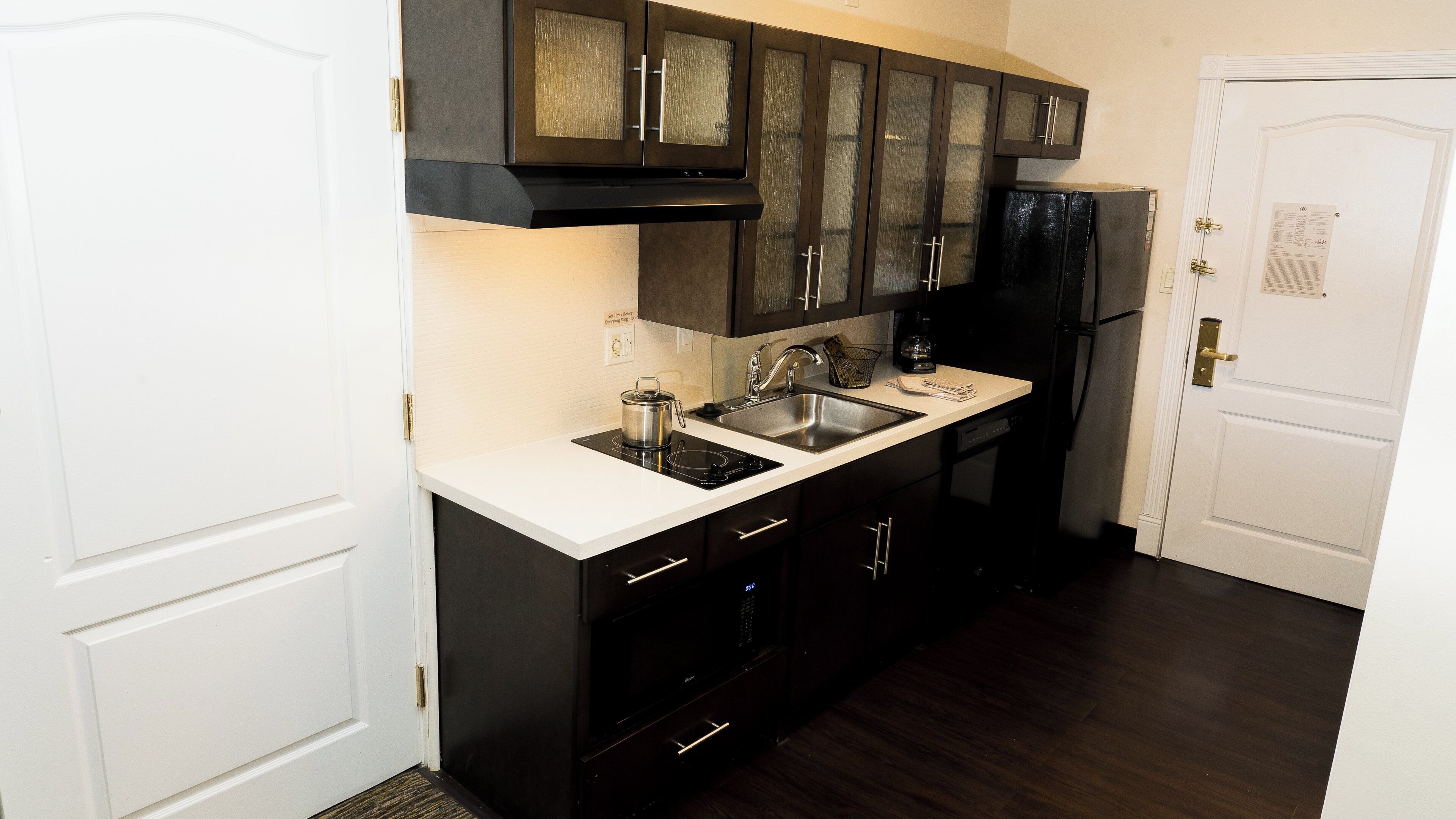 Una cocina con gabinetes oscuros, una encimera blanca, un fregadero de acero inoxidable y un refrigerador negro.