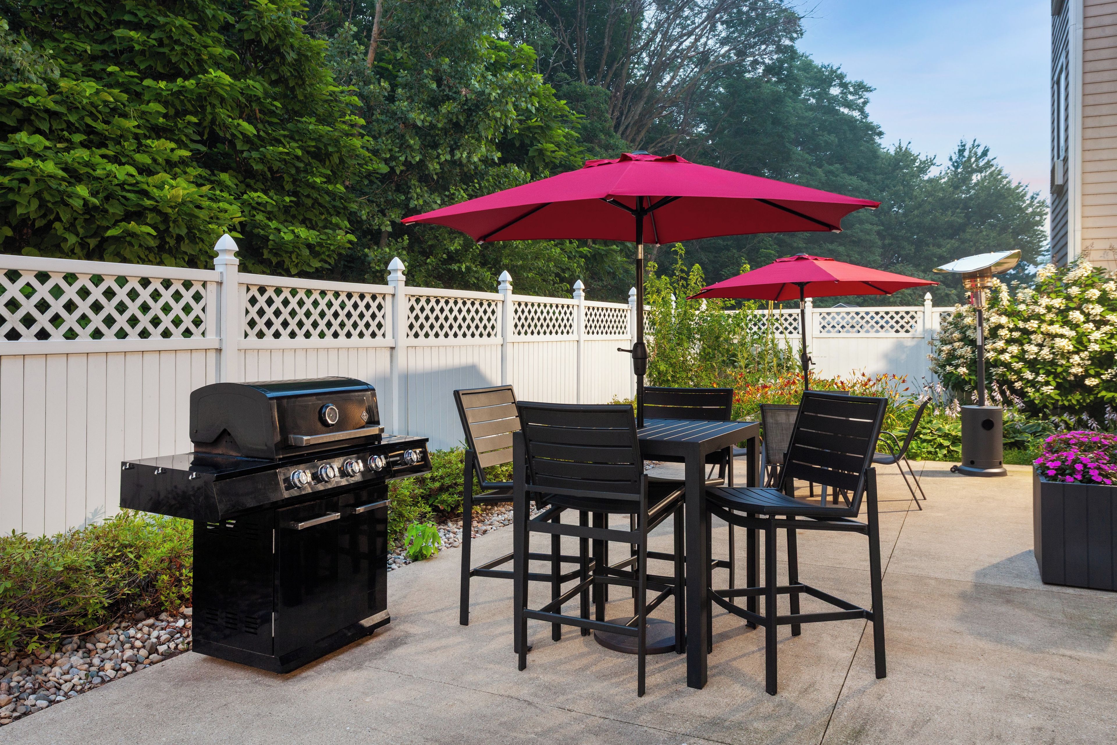 An outdoor patio with a grill, dining table, chairs, and red umbrellas.