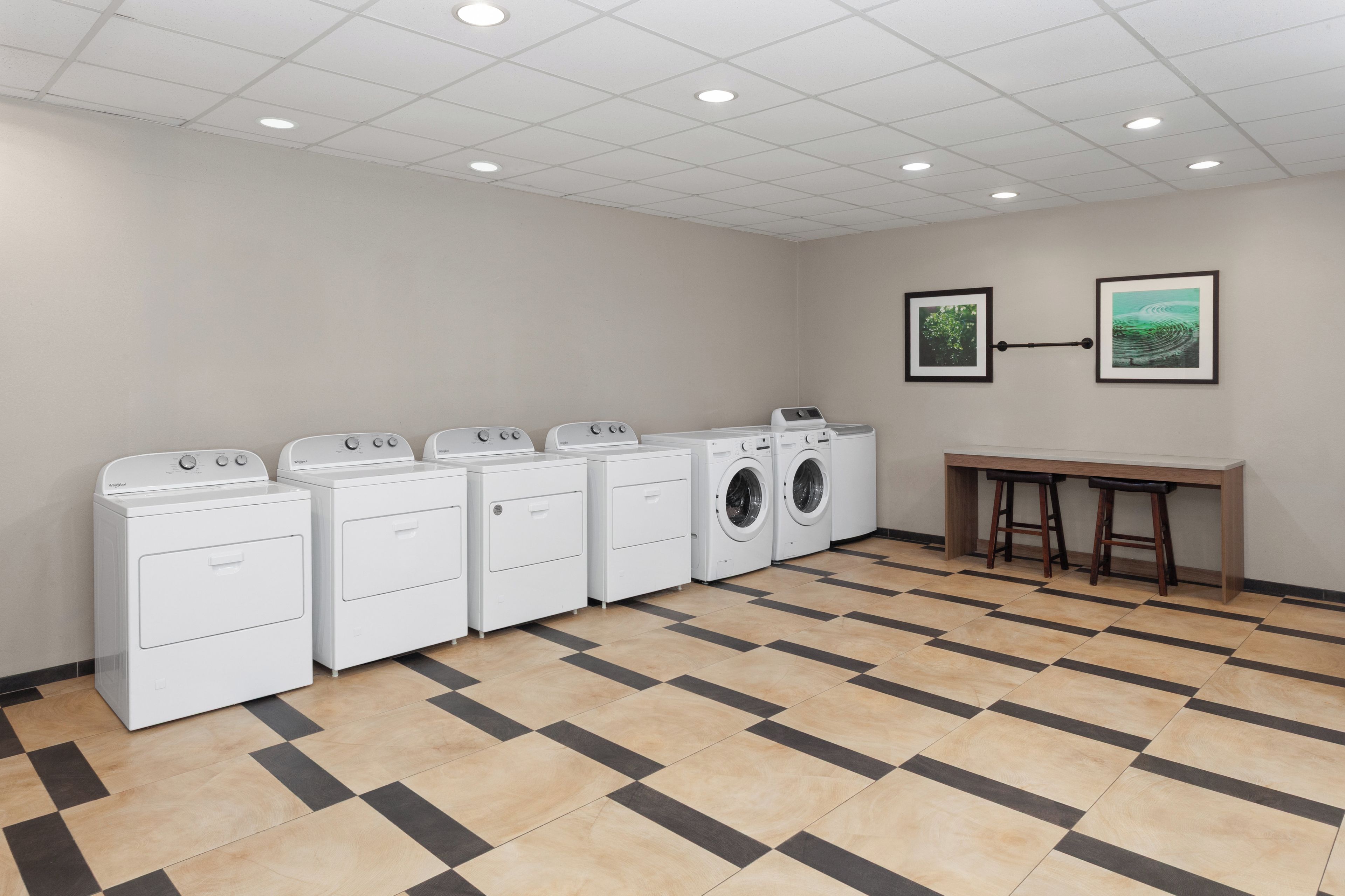 Une buanderie équipée de cinq machines à laver et de cinq sèche-linge blancs, une table en bois avec deux chaises, et deux tableaux encadrés accrochés au mur.