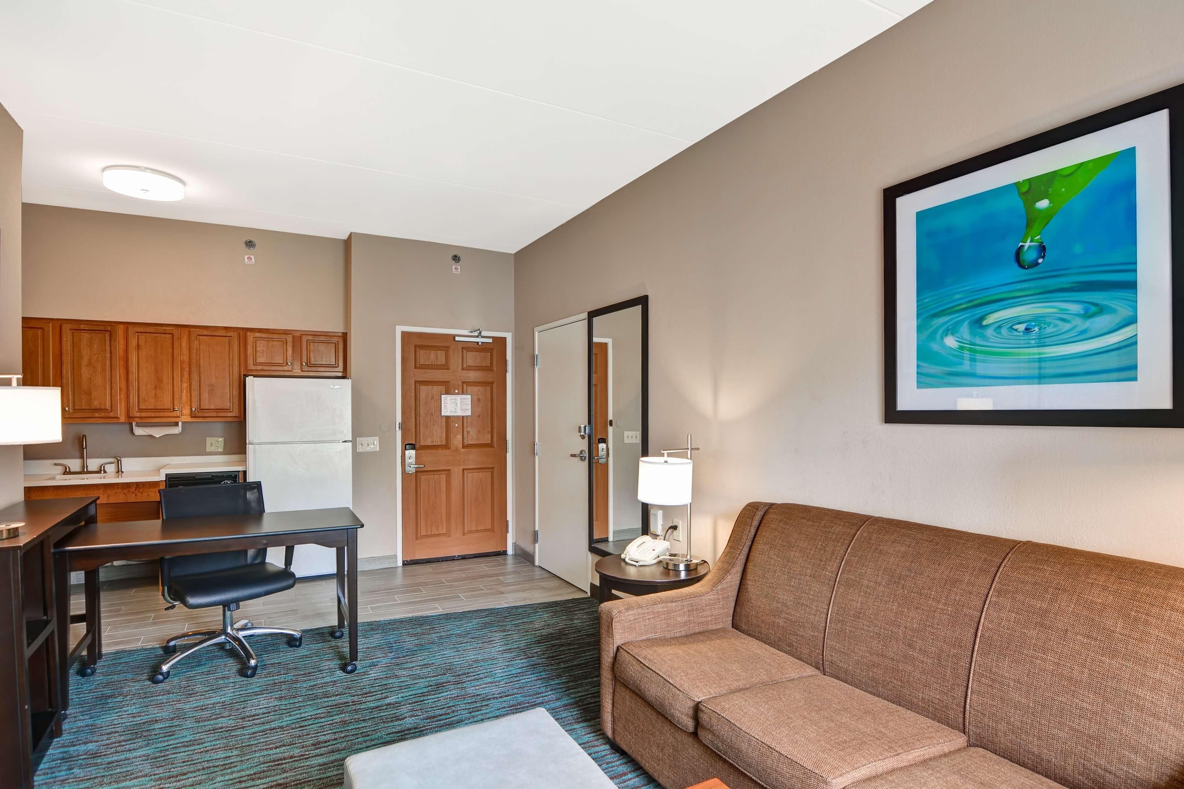A hotel room with a kitchenette, a sofa, a desk with a chair, a framed picture on the wall, and a small table with a lamp.