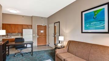 A hotel room with a kitchenette, a sofa, a desk with a chair, a framed picture on the wall, and a small table with a lamp.