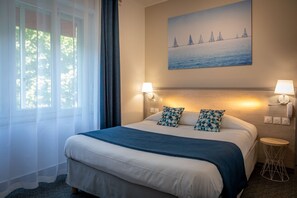 A hotel room with a large bed, two wall-mounted lamps, a painting of sailboats, and a round side table.