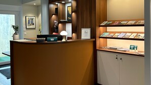 A reception area with a curved counter, framed artwork, a desk with a lamp, and a display of brochures. - Rex Hotel (Lorient)