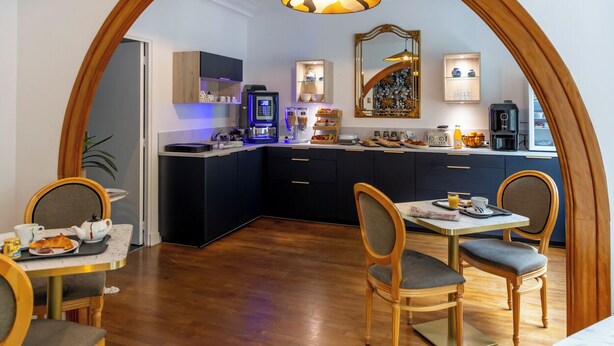 A modern kitchen with dark cabinetry, a coffee machine, and a dining area with a table and chairs. - Rex Hotel (Lorient)