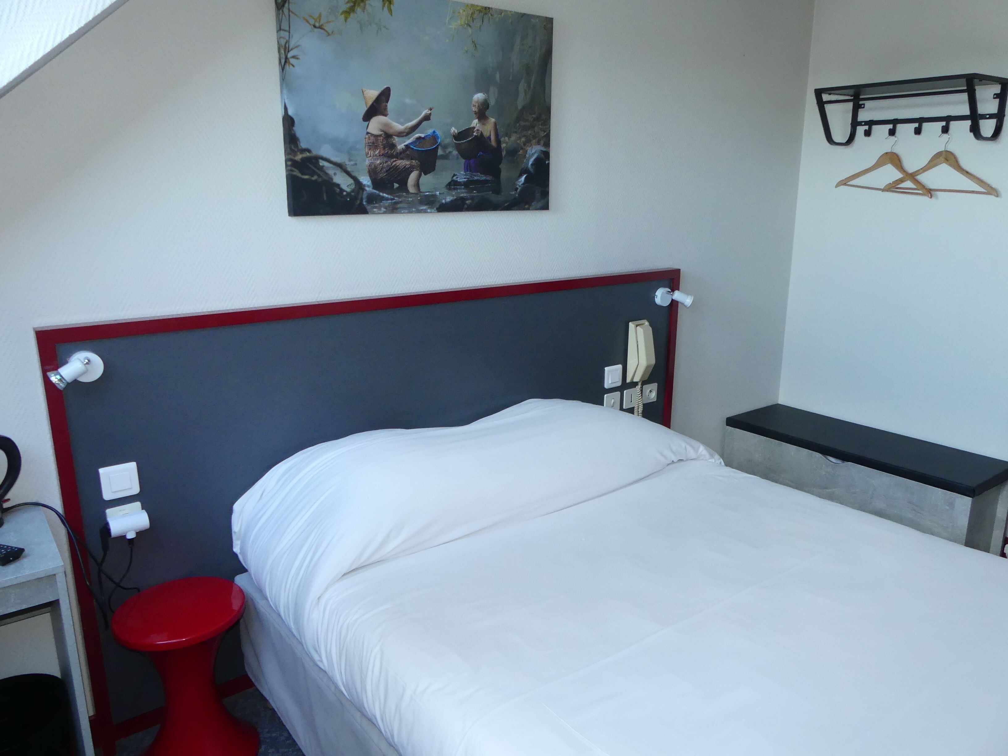 Una habitación de hotel con una cama, un taburete rojo, un teléfono y un cuadro en la pared.