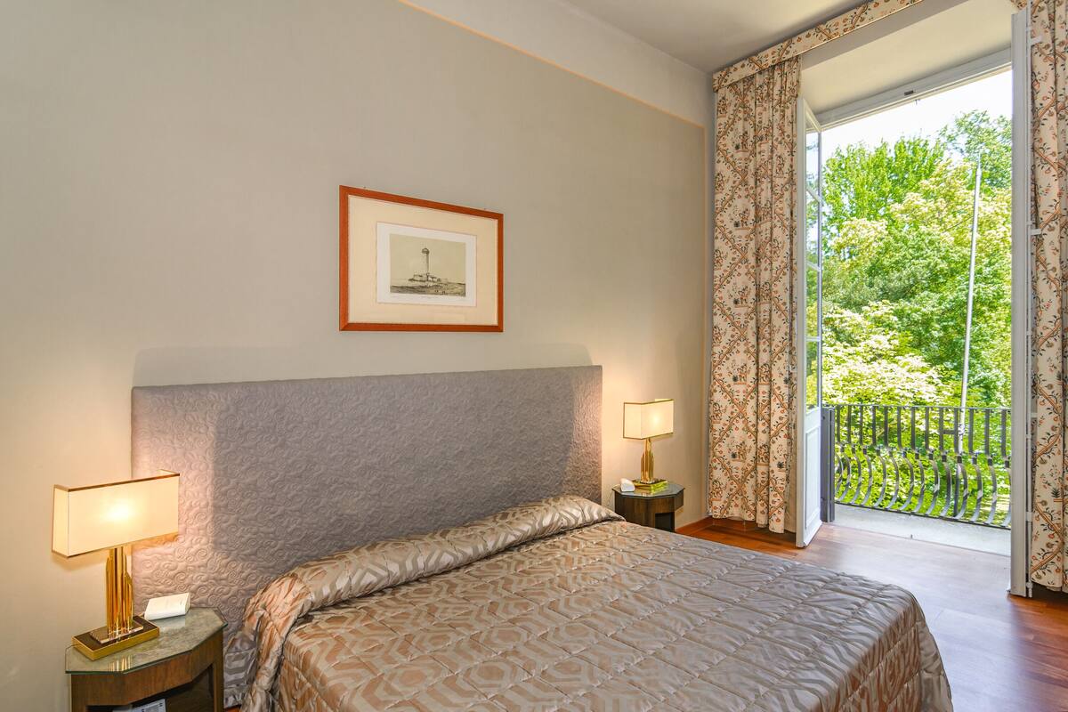 A hotel room with a large bed, two bedside lamps, a framed picture on the wall, and a balcony with a view of greenery.