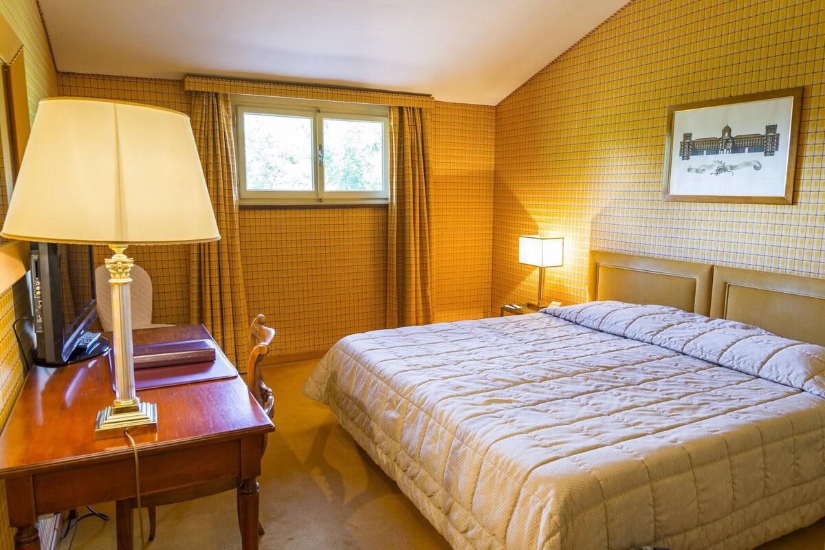 A hotel room with a bed, a desk with a lamp, a chair, a window with curtains, and a framed picture on the wall.