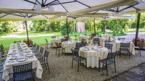 Un área de comedor al aire libre con mesas y sillas redondas, cubierta por sombrillas, sobre un suelo de grava con vista a un jardín.