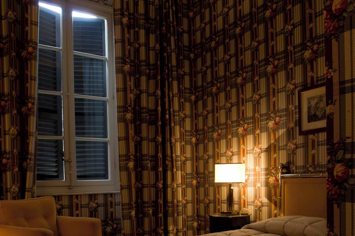A bedroom with a large bed, a chair, a nightstand with a lamp, and a window with blinds.