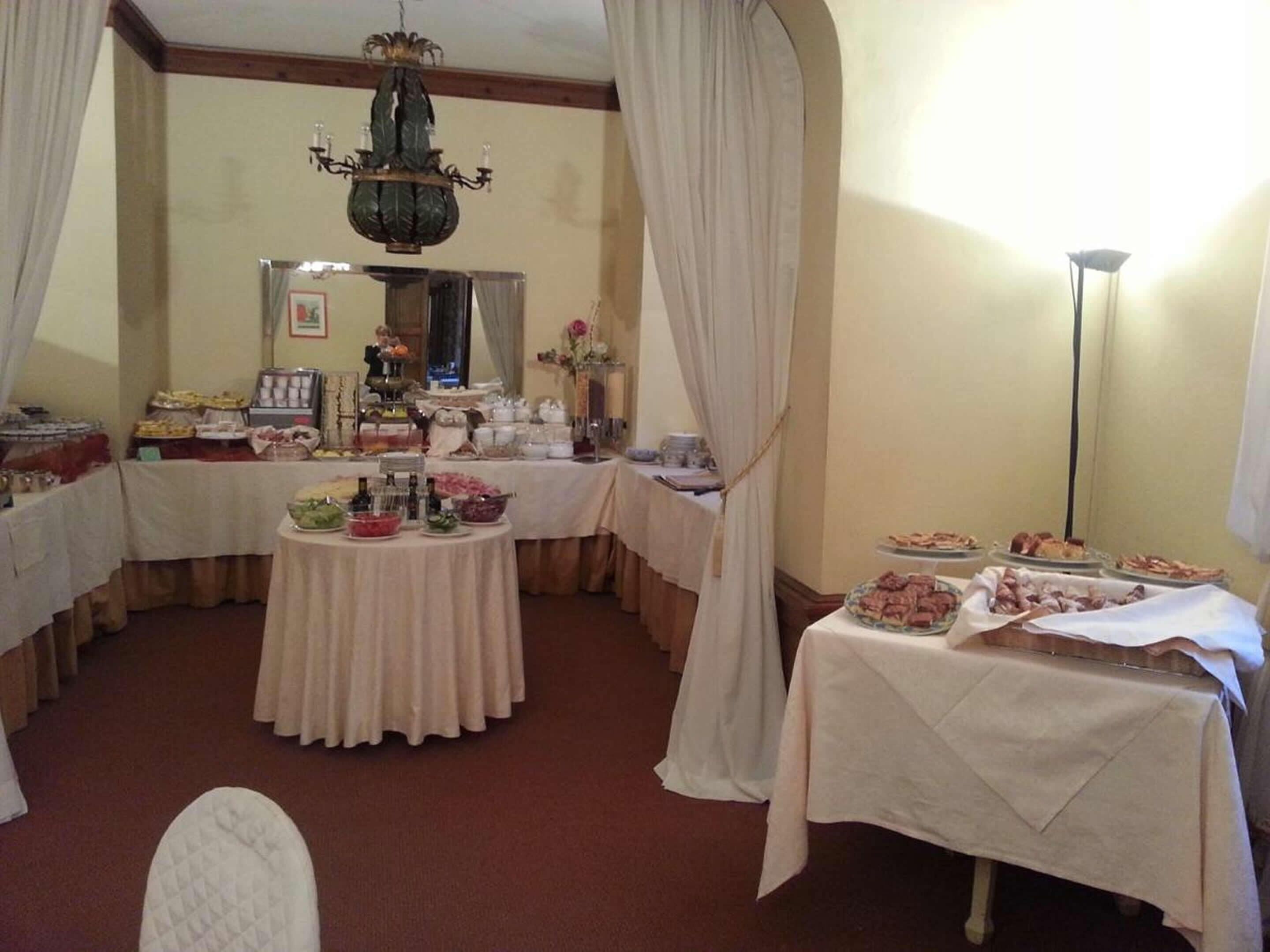 Una habitación con una mesa redonda llena de diversos alimentos, un candelabro y cortinas con drapeados.