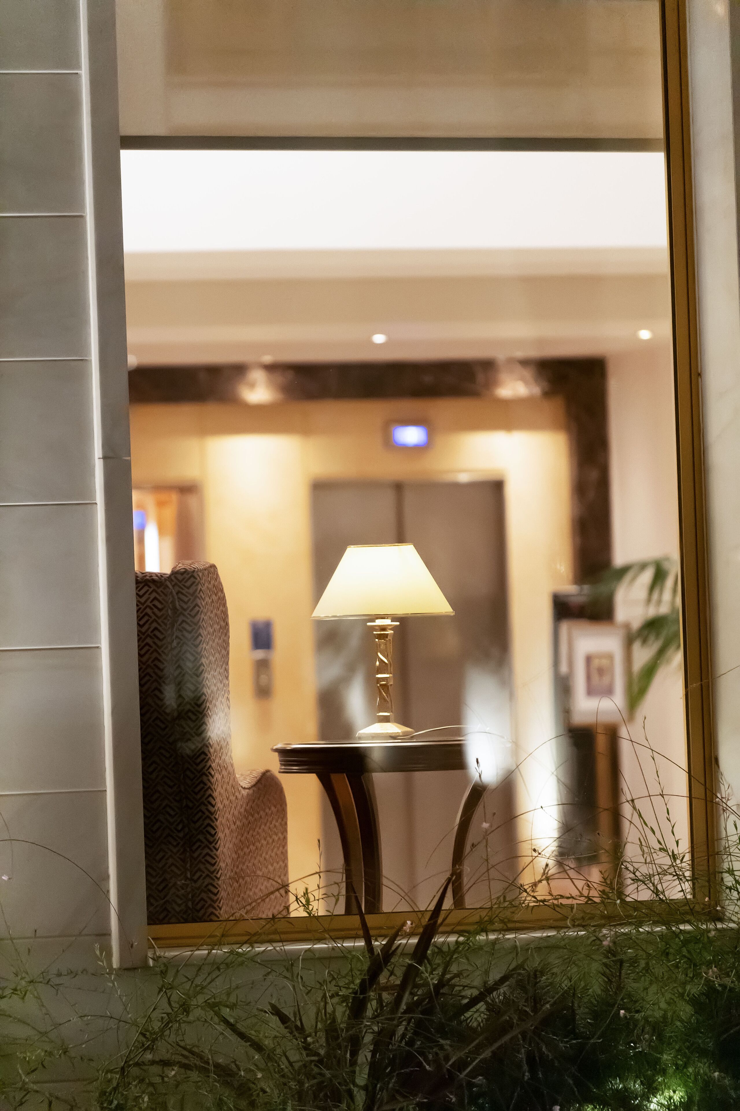 A view through a window with a lamp on a table, a chair, and a door in the background.