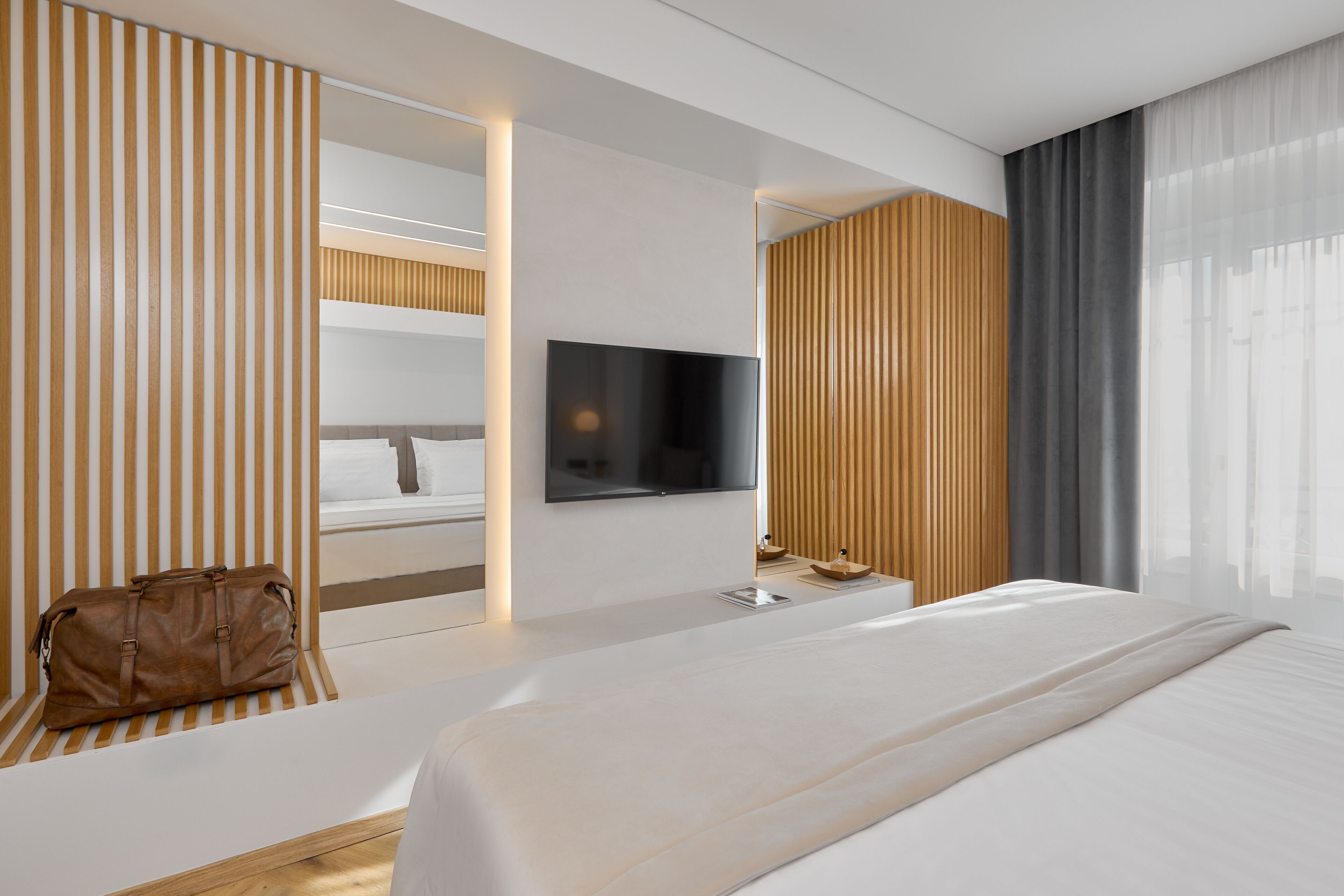 Habitación de hotel moderna con una cama grande, un televisor de pantalla plana montado en la pared, una mesita de noche de madera y un bolso de cuero marrón sobre la cama.