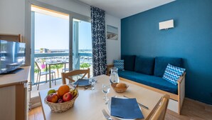 A room with a balcony, a dining table with fruits and glasses, a sofa with cushions, and a television on a stand. - Odalys Residence du port (Hourtin)