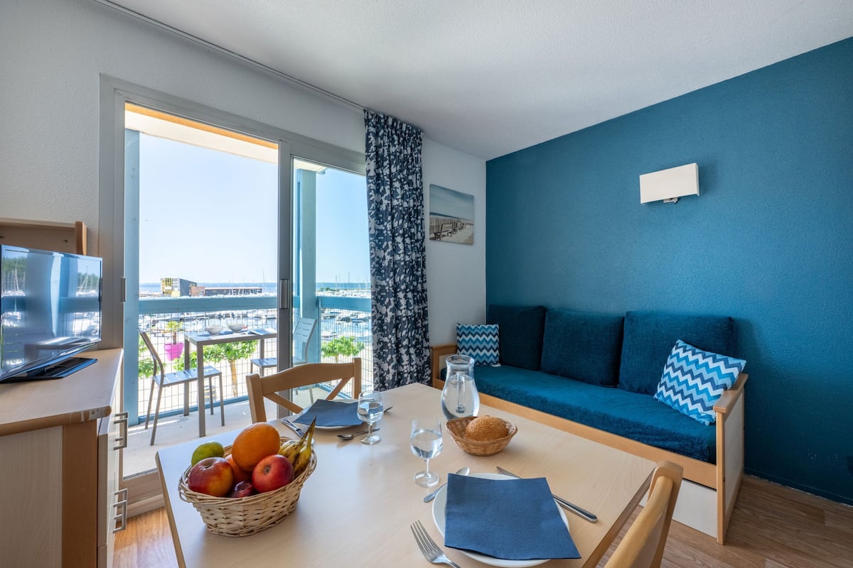 A room with a balcony, a dining table with fruits and glasses, a sofa with cushions, and a television on a stand.