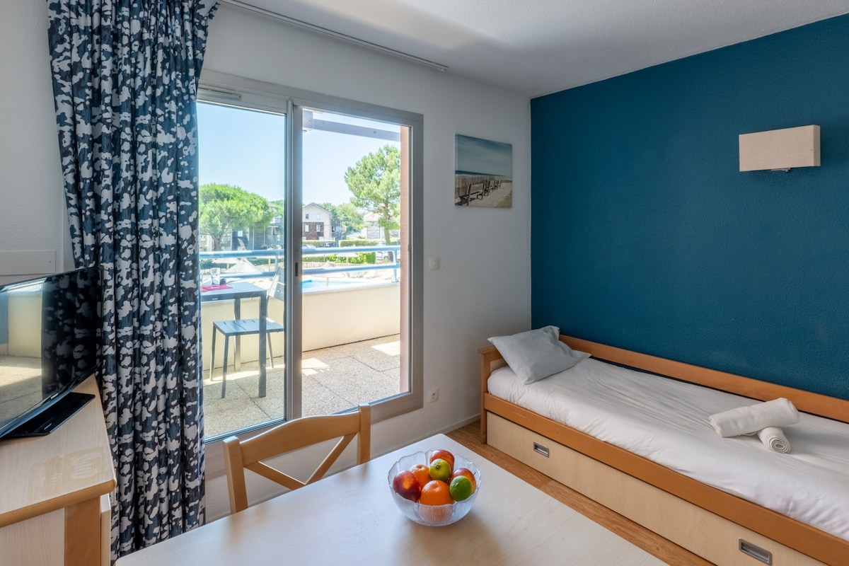 A room with a bed, a desk with a laptop, a bowl of fruit, and a view of the outside.
