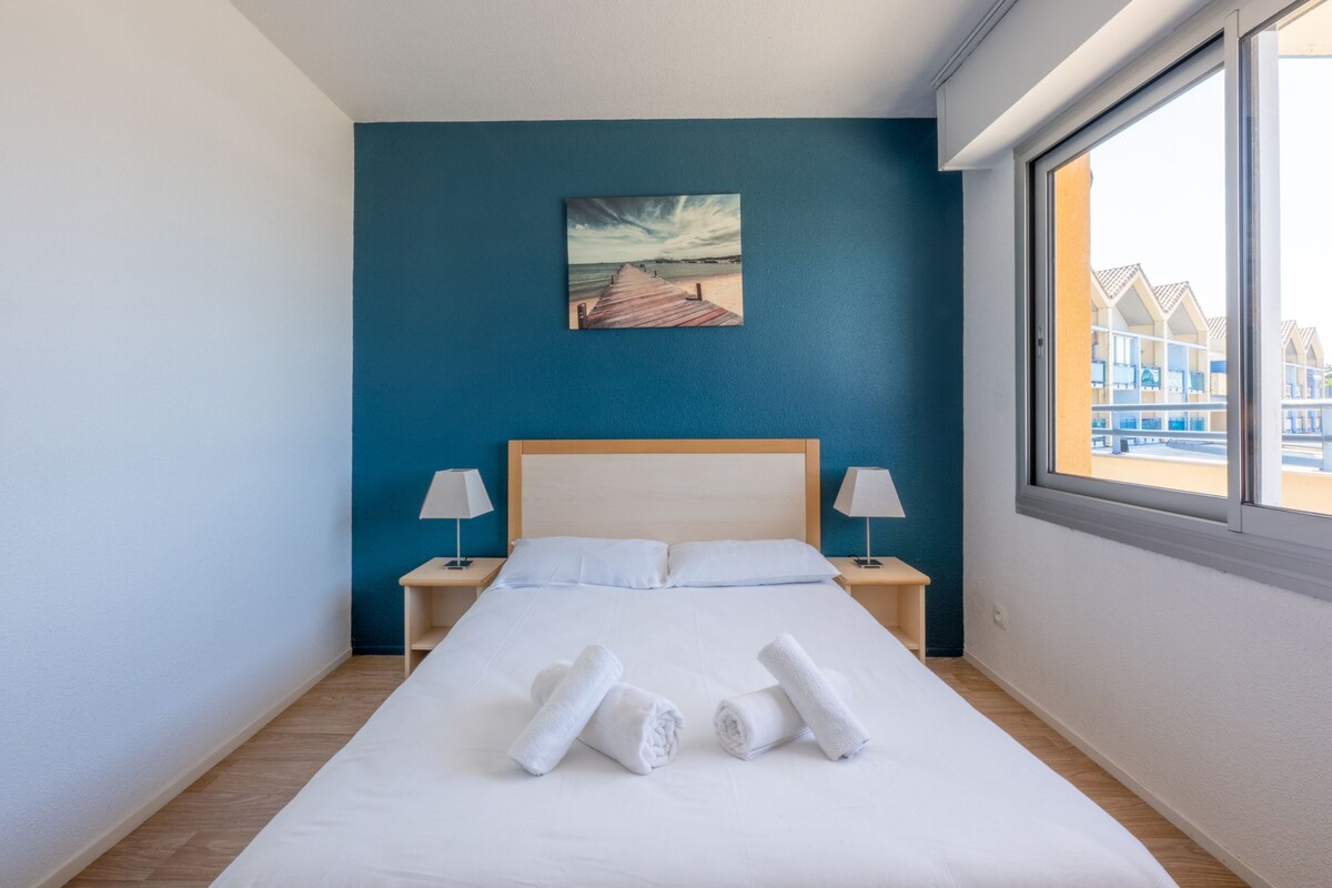 A neatly made bed with white linens, two bedside lamps, a painting of a beach scene, and a window with a view of buildings.