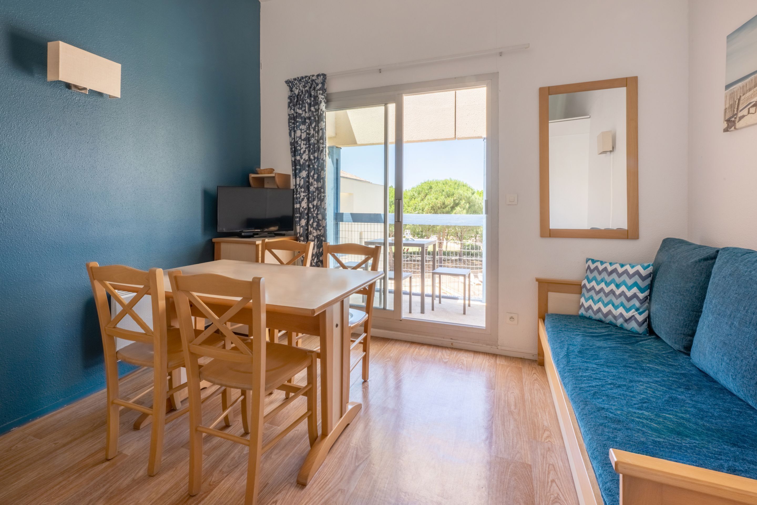 A room with a wooden dining table and chairs, a sofa, a TV, and a balcony with a view of greenery.