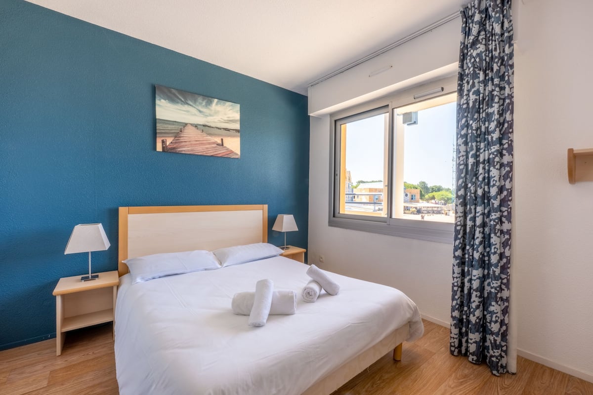 A neatly made bed with white linens and two rolled towels, a painting of a beach scene on the wall, a window with a view of buildings, and a curtain with a floral pattern.