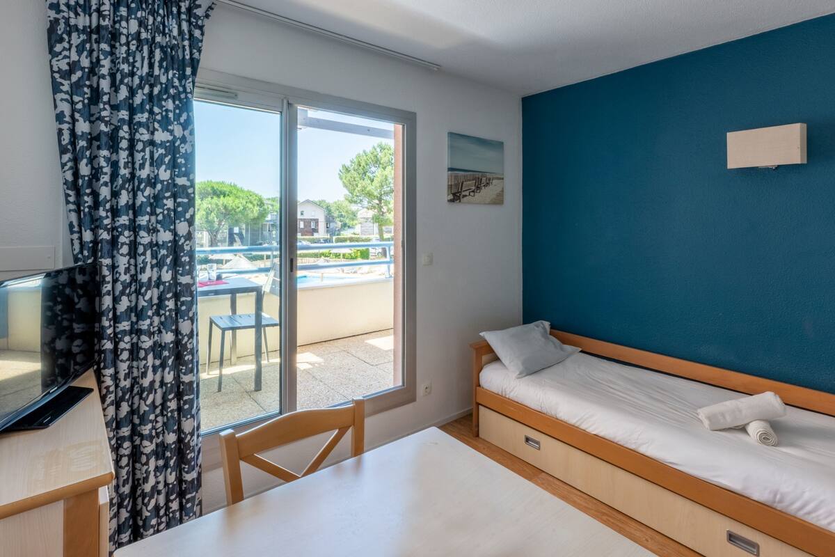 A room with a bed, a desk with a laptop, a window with curtains, and a view of the outside.
