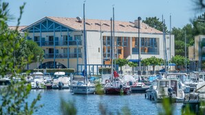 A marina with various boats docked, a building with blue and white exterior, and a clear sky. - Odalys Residence du port (Hourtin)