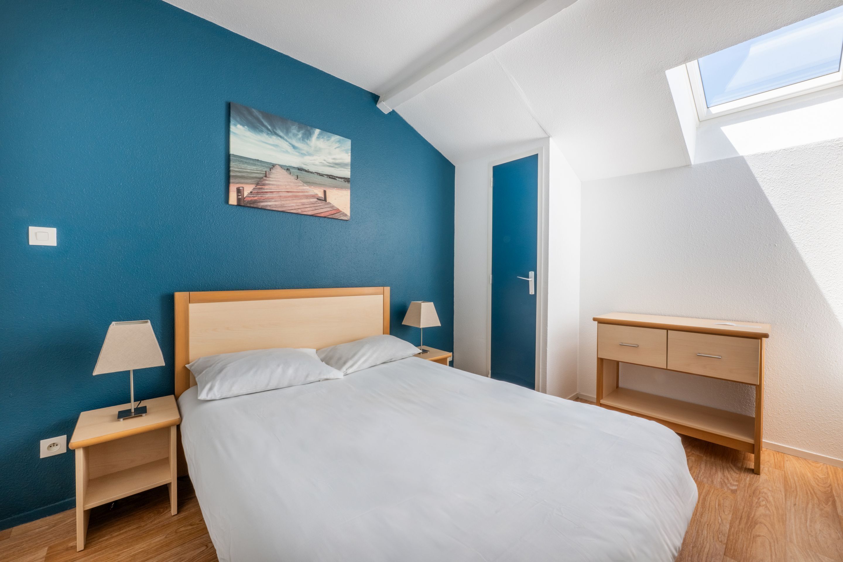 Un dormitorio con cama, mesitas de noche, un escritorio y un cuadro en la pared.