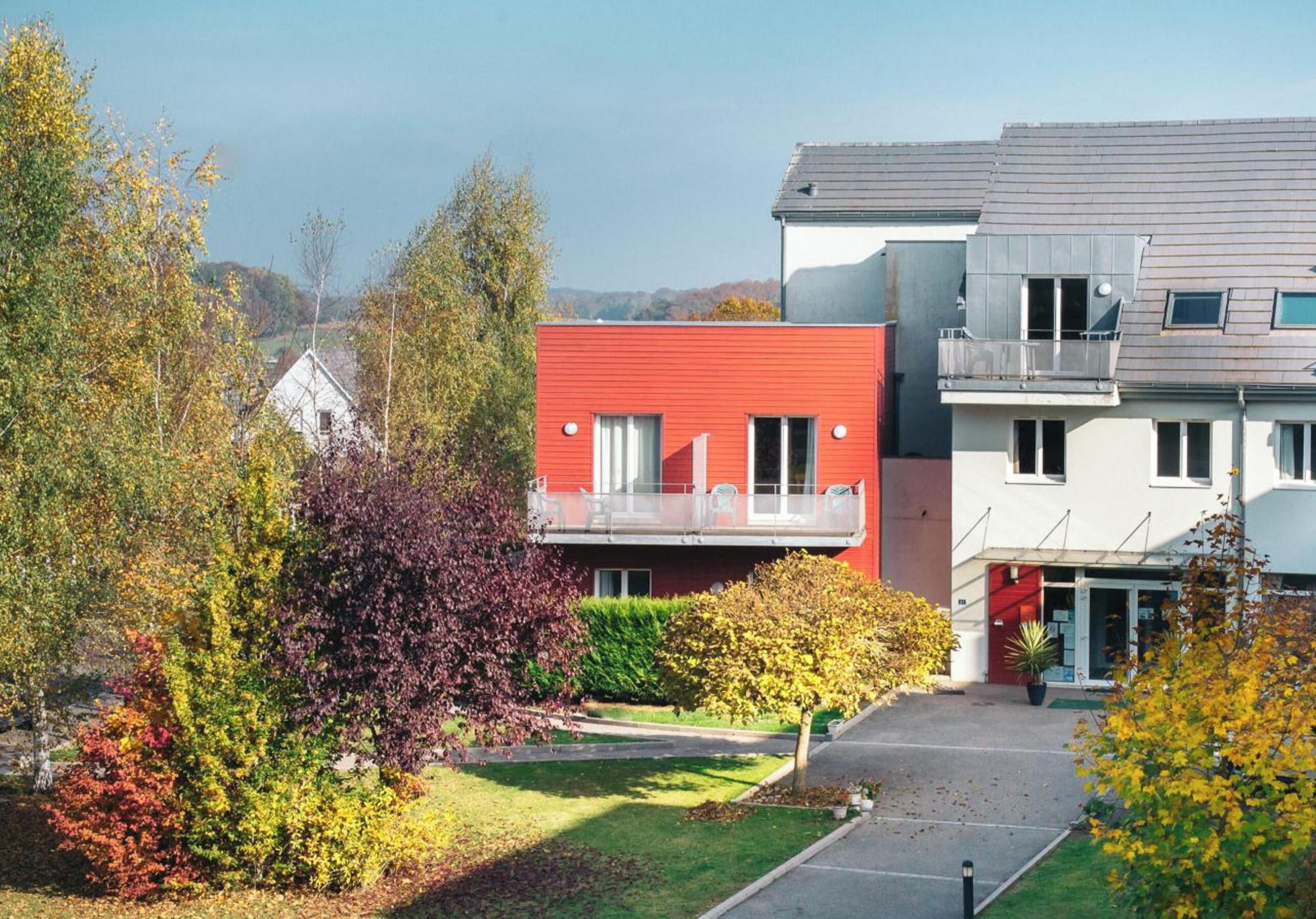 Ein modernes Wohngebäude mit roter Fassade, einem Balkon und einem Parkplatz.
